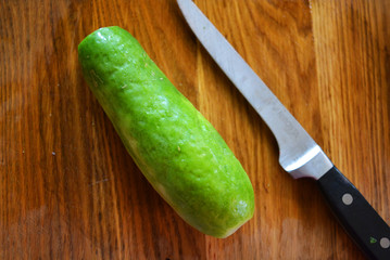One big green fresh cucumber with a metal knife on a brown wooden kitchen board.