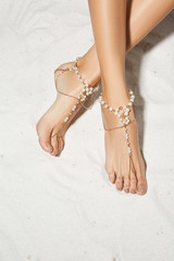 Cropped top view shot of lady's crossed legs on the sandy background. The girl with light brown pedicure is wearing a golden anklet adorned with chains and pearls. Trendy women's summer accessory.