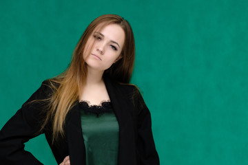 Fototapeta premium Concept close-up portrait of a pretty girl, a young woman with long beautiful brown hair and in a black jacket on a green background. In the studio in different poses showing emotions.