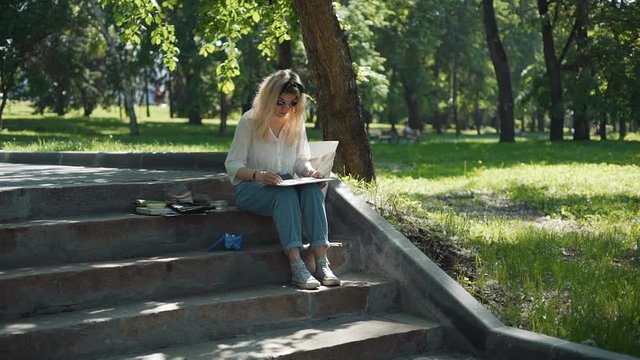 Female Street Artist Doing a Sketch for Future Picture Outdoors in Slow Motion. Professional Painter Working in a City Park in Sunny Summer Day while Sitting on Steps.