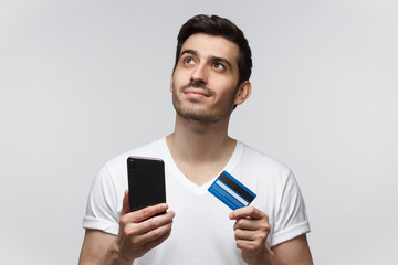 Young handsome man thinking about online shopping via internet, holding credit card and smartphone...