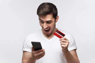 Young happy man holding smart phone and credit card in hands, looking at screen with amazement,...