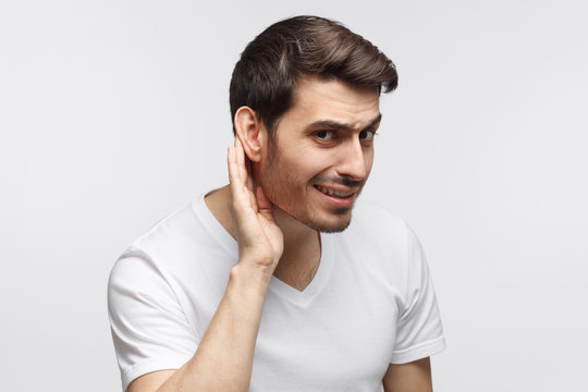 Young man pressing hand to ear trying to hear something better isolated on gray background