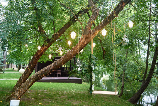 Wedding swing with orchid flowers and monstera palm leaves hanging on tree decorated with lanterns and garland, copy space. Wedding decorations - Powered by Adobe