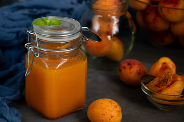 Apricot jam in a transparent jar, near the whole fresh fruit, spread on bread dessert. All on a gray, stone background.
