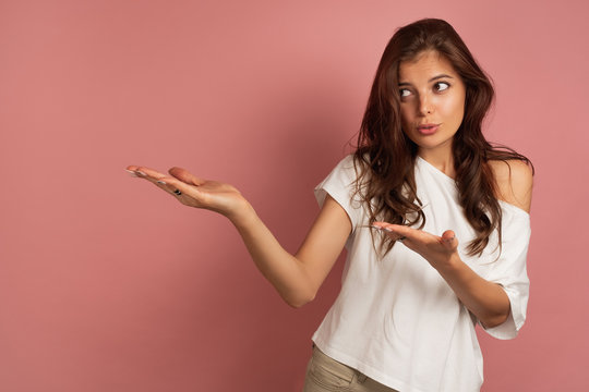 Young brunette points at the left space at the picture with a question in her eyes, pink background
