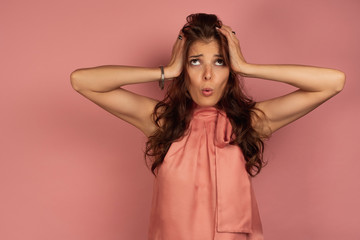 Fototapeta premium Young brunette holds her head in perplexity with a funny face expression, pink background