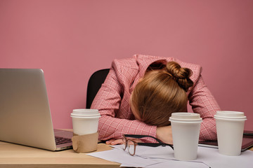 Young woman lays at the table because she is tired from job, pink background