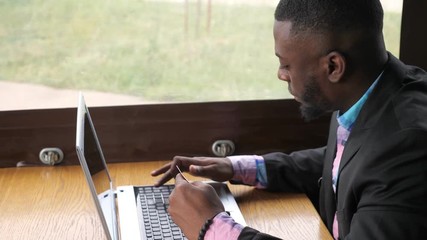 Black man makes payment bank card on laptop. Online shopping sitting in cafe. Young afro american businessman is buying online entering his data to computer from credit card, side view. Guy customer.
