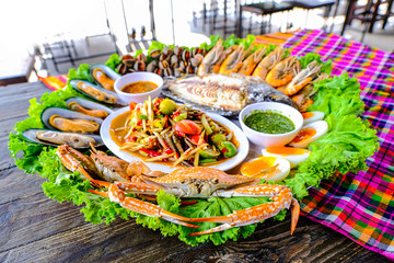 Seafood Somtum has clams, shrimp, crabs, boiled eggs, grilled tilapia, placed in a beautifully placed tray on a wooden table.