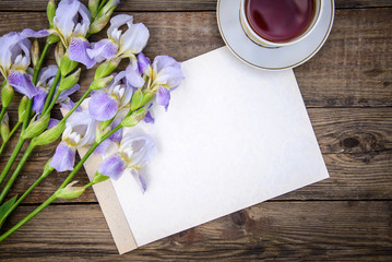 Beautiful purple flowers irises, a sheet of paper and a cup of tea on a wooden background