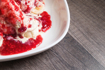 Closeup Vanilla ice cream poured with strawberry or raspberry jam with sliced banana slices on a white plate on a dark wooden table. You can do this at home on vacation