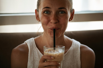 Beautiful thoughtful smiling girl drinking a coffee beverage through a straw sitting in the cafe near the window - Powered by Adobe