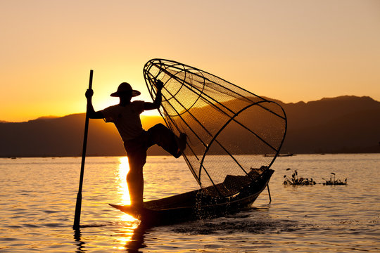 Silhouette of Myanmar fisherman on wooden boat at sunset .Burmese fisherman on bamboo boat catching fish in traditional way with handmade net balancing and paddling with a leg. Inle lake,Myanmar