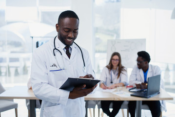Handsome Afro American doctor in white coat makes some notes. Young medical student with a stethoscope around his neck holds a folder.
