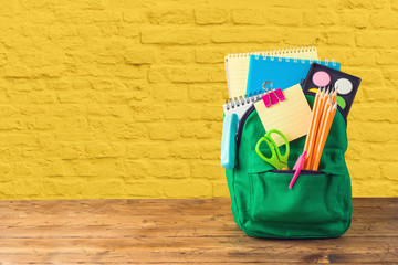 Back to school concept with bag backpack and school supplies on wooden table over yellow brick wall...