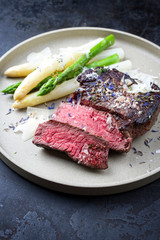 Barbecue dry aged wagyu sirloin beef steak sliced with green and white asparagus and parmesan cheese as closeup on a beige modern design plate