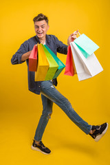 Impressed young man with colorful shopping bags in hands with open mouth, happy after successful shoping, isolated over yellow background