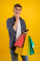 Handsome guy with shopping bags is using a mobile phone and smiling. Portrait of a smiling man holding shopping bag over yellow background and looking at camera