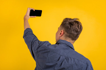 Attractive young man with smartphone on color yellow background. Cheerful young man wearing plaid shirt standing isolated over orange background, taking a selfie