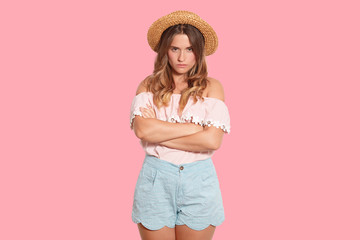 Attractive upset female with frowned face standing over pink background in studio, having folded arms, looking directly at camera, being disappointed, wearing blouse, blue shorts and straw hat.