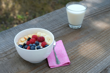 Close up a white bowl of rolled oats served with milk, slices of bananas, fresh apple, wild strawberry, blue berries, red raspberries and blackberries. Healthy breakfast outside in summer.