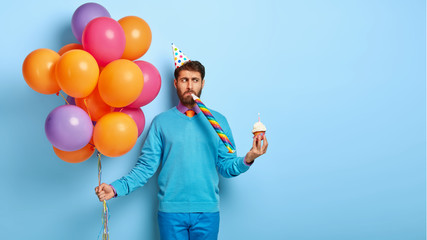 Handsome young man blows party horn, wears cone hat, holds air balloons, small muffin, being angry...