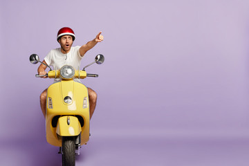 Scared man drives fast yellow scooter, wears protective helmet and white t shirt, points index finger in distance, notices scarying scene on road, poses against purple wall, blank copy space. Driving