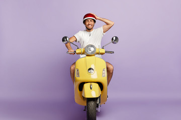 Embarrassed young guy clenches teeth, keeps hand on helmet, poses on yellow retro motorbike, forgets about driving licence, has good driving skills, enjoys free time, isolated on purple wall