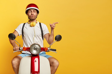 Funny unshaven guy in casual wear, drives on motorcycle, has good trip, enjoys freedom, points index finger on blank space over yellow wall, pouts lips. Professional driver bought new scooter - obrazy, fototapety, plakaty