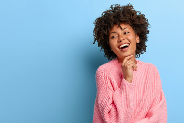 Fototapeta na wymiar Charismatic beautiful young Afro woman touches chin gently, smiles broadly and shows white teeth, looks aside, enjoys friendly company, wears loose rosy sweater, models against blue background.