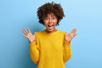 Photo of emotional curly haired young woman exclaims with joy, raises palms, dressed in yellow...