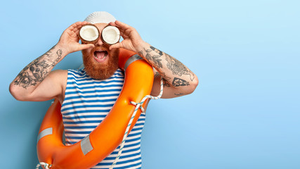 Summer lifestyle concept. Bearded curious male sailor wears swimhat, striped vest, keeps halfs of...