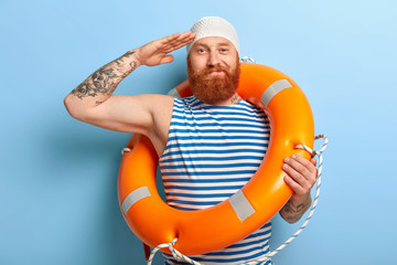 Satisfied ginger man makes salute soldier gesture, wears white swimhat, sailor striped vest,...