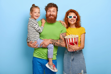 Merry ginger family have friendly relationship, spend free time all together, husband and wife visit movie with daughter, watch cartoon, woman wears three dimensional glasses, holds popcorn.