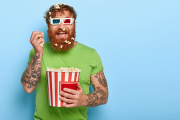 Glad cheerful man has fun, eats popcorn in cinema, enjoys funny movie, good company, wears 3d glasses, green t shirt, has tattooed arm, stands against blue wall with blank space. Fast food in theatre - obrazy, fototapety, plakaty