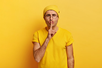 Serious man with mysterious look, keeps index finger over lips, looks straightly at camera, demands complete silence, makes hush sign, wears big roud glasses, yellow clothes. Secrecy concept