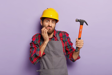 Home renovation concept. Indignant builder holds hammer, does repairing, wears building helmet, has...