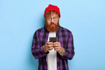 Fashionable hipster with red hair and thick beard, focused in smart phone, recieves link to some...