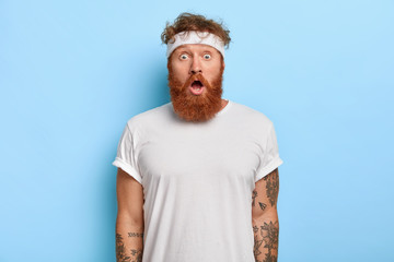 Indoor shot of surprised bearded man with red hair, stares with astonishment, has gymnastic every...