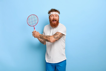 Emotive active adult man with ginger hair holds badminton racket, gazes happily at camera, likes game, dressed in white t shirt and blue shorts, poses indoor. People, leisure and sport concept