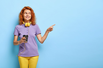 Positive teenage girl with red short hair, points away on free space, holds paper cup of coffee, has joyful smile, enjoys hot drink, wears casual outfit, makes choice and decides what to buy