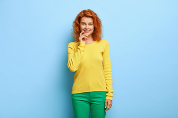 Photo of glad satisfied woman keeps index finger near lips, has wavy ginger hair, enjoys pleasant talk with interlocutor, wears yellow jumper and green pants, isolated on blue background. Emotions