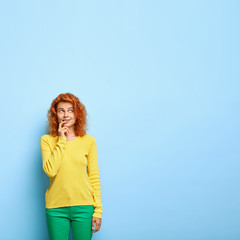 Thoughtful red haired lady keeps finger on lips, thinks about something, focused upwards, wears bright clothes, poses against blue wall, empty space for your promotion. Pensive millennial girl