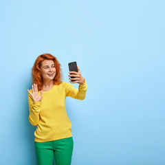 Lovely redhead woman makes video call, talks with relatives, waves at camera, has friendly smile, greets someone on distance, wears bright outfit, poses indoor against blue background, free space