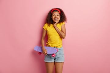 Satisfied cute Afro woman has motor activity, holds skateboard, feels relaxed and delighted, wears red headband, yellow t shirt and jean shorts, enjoys summer vacation and recreation, stands indoor