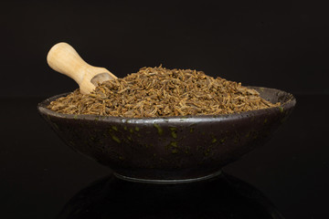 Lot of whole dry caraway fruits in a ceramic bowl with wooden scoop in it isolated on black glass