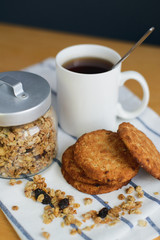 round brown wholemeal oatmeal cookies stack with granola and black tea in white cup on striped textile napkin on wooden table, close up side view of vertical still life stock photo image