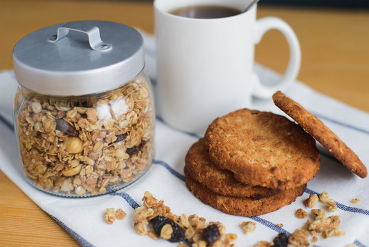round brown wholemeal oatmeal cookies stack with granola and black tea in white cup on striped textile napkin on wooden table, close up side view of horizontal still life stock photo image