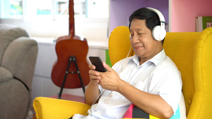 Senior asian man using smartphone   listening music with headphones, sitting on sofa in living room...
